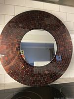 Front view of round wall mirror showing mosaic tile frame in reddish-brown tones and clear central mirror glass.