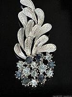 Close-up of the lapel pin showing textured silver tone metal leaves and clustered dark and light blue stones.