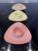 Three Melmac egg cups arranged in line on a black stovetop surface, colors pink, yellow, and tan visible with atomic retro shape.