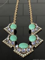 Front view of gold-tone chain necklace with geometric deco-style pendant, turquoise-colored stones, and light purple crystals.