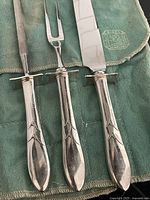 Close-up view of the silver carving knife, fork, and sharpening steel handles showing the polished finish and design details, resting on the green Birks storage bag.