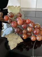 Front view of large grape cluster with polished carnelian stones in varying sizes attached to metal wire stems and natural stone carved leaves, showing the piece set on a reflective surface.
