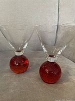 Two mid-century modern martini glasses with clear bowl and red bubble ball stems set on a beige fabric background.