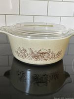 Side view of vintage Pyrex Forest Fancies casserole dish with mushroom pattern and clear lid on black stovetop.
