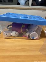 Top-down view of purple bathtub, white dog figure, two white teacups with faces in transparent bin with blue lid.