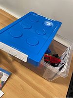 Clear plastic bin with blue lid on floor containing Lego parts and manuals inside
