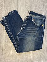 Blue men's True Religion jeans folded on floor, showing classic contrast stitching and faded wash details on thighs.