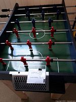 Top view of foosball table showing red and blue player figures placed on metal rods, green playing surface with white markings and wooden legs.