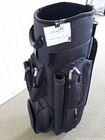 Black and brown Top Flite golf bag standing upright on carpet with multiple zippered compartments and a padded strap, showing the top open tube sections for golf clubs.