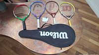 Four junior tennis racquets laid out side by side on table with Wilson black zippered carrying case beneath them