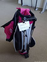 Photo showing ultra light kids golf bag in pink and black with a stand and multiple zippered pockets, tag attached indicating lot number.