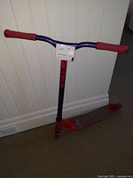 Red and blue kick scooter leaning against wall showing handlebar and deck with red grips and wear marks.