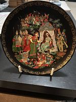 Front of the round decorative plate showing elaborate multi-character scene with a royal figure and attendants, set on a black background with gold ornamental border.