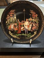 Front view of decorative plate showing detailed folkloric scene with priest and servant Balda on black background with gold rim designs.