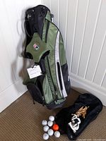 Green and black senior golf bag standing upright against a white paneled wall, with eight golf balls, wooden tees, a white golf glove, and a black shoe bag placed on a carpeted floor in front of it.