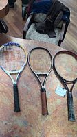 Three senior tennis racquets displayed on a table: one Wilson with blue frame, one Head with black frame and wooden grip, one Yonex with black and red frame