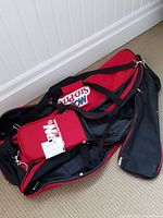 Photo of a red and black SloPitch National sports bag placed on a carpeted floor against a white paneled wall, showing the main compartments and adjustable shoulder straps.