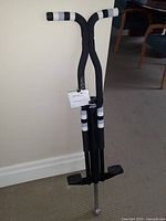 Photo of the New Bounce Sport Pro Pogo Stick showing its black and white design with padded grips and footrests, standing upright on a carpeted floor against a wall.
