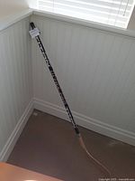 Photo shows the full Titan Ultra Pro hockey stick standing upright against a white paneled wall and a window with blinds, highlighting the black shaft with branding and the wooden blade.