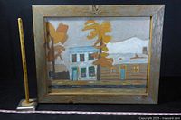 Photo of the full painting showing the folk-art style scene of buildings and trees in barn-wood frame, with measurement sticks for scale.