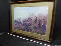 Angled view of the large framed print showing three horseback riders in hunting attire with hunting dogs in a wooded area. The green mat and wooden frame with bamboo-style corners are visible.