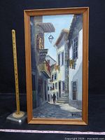 Framed oil painting of a narrow cobblestone Spanish street with buildings, lamp, stairs, and walking figure.