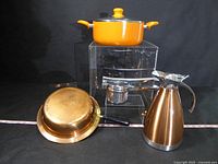 Four kitchen items on clear acrylic risers: copper saucepan, orange pot, chrome juicer, and copper coffee pot