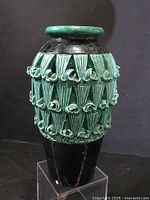 Full view of the tall handmade ceramic vase with green glazed top rim and raised decorative patterns on the dark base.