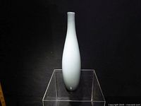 Single tall white hand blown glass vase photographed on a clear display stand against a dark background.