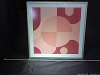 Full view of the large framed abstract print under glass with a silver frame, geometric shapes in pink and red.