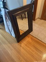 Mirror leaning against wall on hardwood and tile floor, showing front view with detailed metal frame and reflective glass surface.