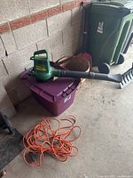 Green electric leaf blower on purple bin with orange extension cord coiled on floor, showing the blower's top and handle