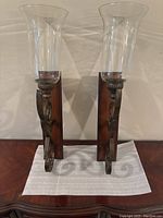Pair of two post iron wall sconces with wooden backplates and tall flared glass hurricane candle holders, viewed frontally.