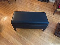Black vinyl upholstered storage bench shown on hardwood floor from above, closed top view.