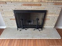 Front view of black iron single-panel fireplace screen with mesh center and decorative top and bottom edges, placed in front of brick fireplace with concrete hearth.