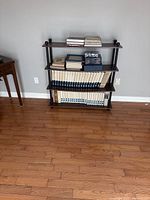 Full view of vintage encyclopedia books and additional older books on a three-tier wooden shelf with metal supports against a gray wall on hardwood floor.