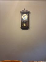 Full view of vintage wooden wall-mounted grandfather clock against beige wall showing clock face and lower pendulum section.