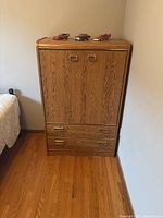 Front view of wooden armoire showing two doors and two drawers with brass-tone handles, natural wood grain finish.