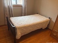 Wooden twin bed frame with mattress and cream-colored quilt in a room with hardwood floor and window with curtains.