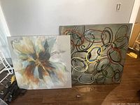 Two abstract paintings leaning against a wall on a wooden floor. The larger one measures 40x40 inches and shows a floral/explosive design in earthy tones.