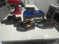 Three pairs of men's Vince Camino shoes displayed on a kitchen counter with two boxes and kitchen appliances in background.