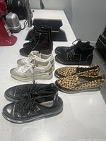 Five pairs of women's shoes including black ankle boots, black high-top sneakers, white metallic sneakers, leopard print flats, and black patent low-top sneakers on white counter surface.