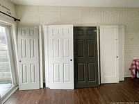 Nine used doors standing indoors against a brick wall, including white and dark gray paneled doors of varying widths and heights.