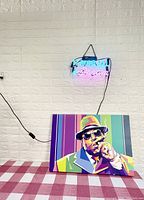 Photo showing colorful Biggie Smalls print sitting on a table with pink checkered cloth below and LED light sign glowing on the wall above with phrase "It was all a dream".