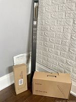 Photo showing boxed Laster LED floor lamp, boxed Laster LED desk lamp, and boxed Umbra drapery rod set leaning against wall