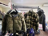 Three jackets hanging on a black metal rack: green Canada Goose jacket on the left, green and black checkered fleece jacket in the middle, black puffer jacket on the right.