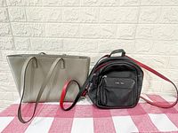 Both the grey DKNY purse and black Calvin Klein backpack displayed side by side against a light brick wall and red and white fabric.