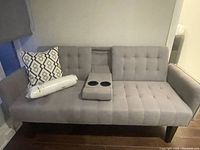 Full front view of gray futon sofa with armrests, tufted upholstery, folded down center console with two cup holders, and two pillows on left.
