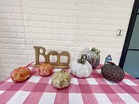 Photo showing five large glass pumpkins in colors orange, gold-speckled, white, and metallic, plus a wooden 'Boo' sign on a red and white checkered tablecloth.