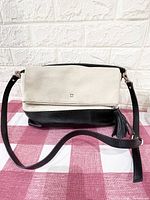 Front view of the purse showing beige fold-over flap with small square metal logo and black leather bottom. Attached black leather adjustable strap.